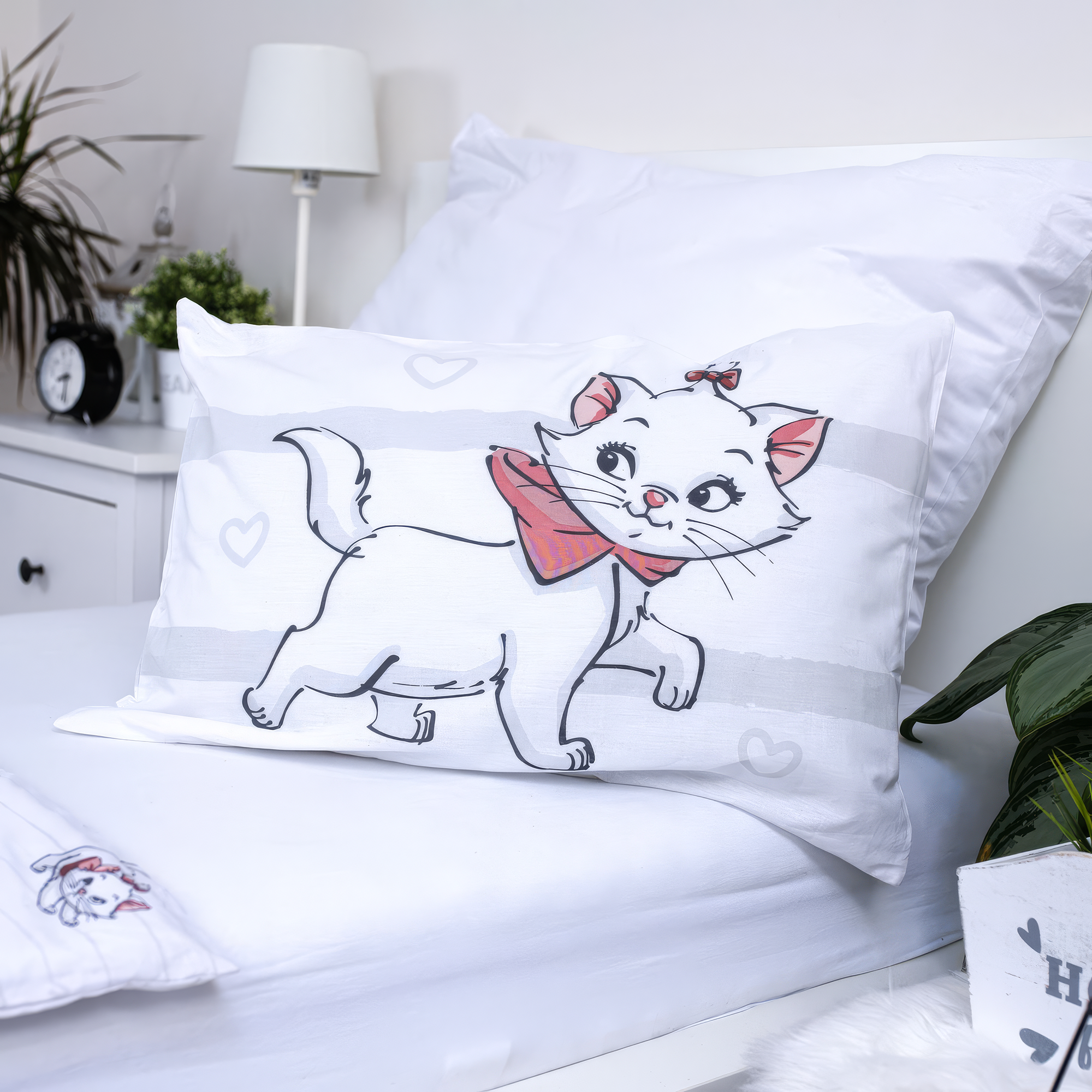 Nahaufnahme des weißen Aristocats-Kopfkissens in einem Bett; zeigt die weiße Katze Marie mit einer rosa Schleife auf einem dezent grau gestreiften Hintergrund mit kleinen Herzen.