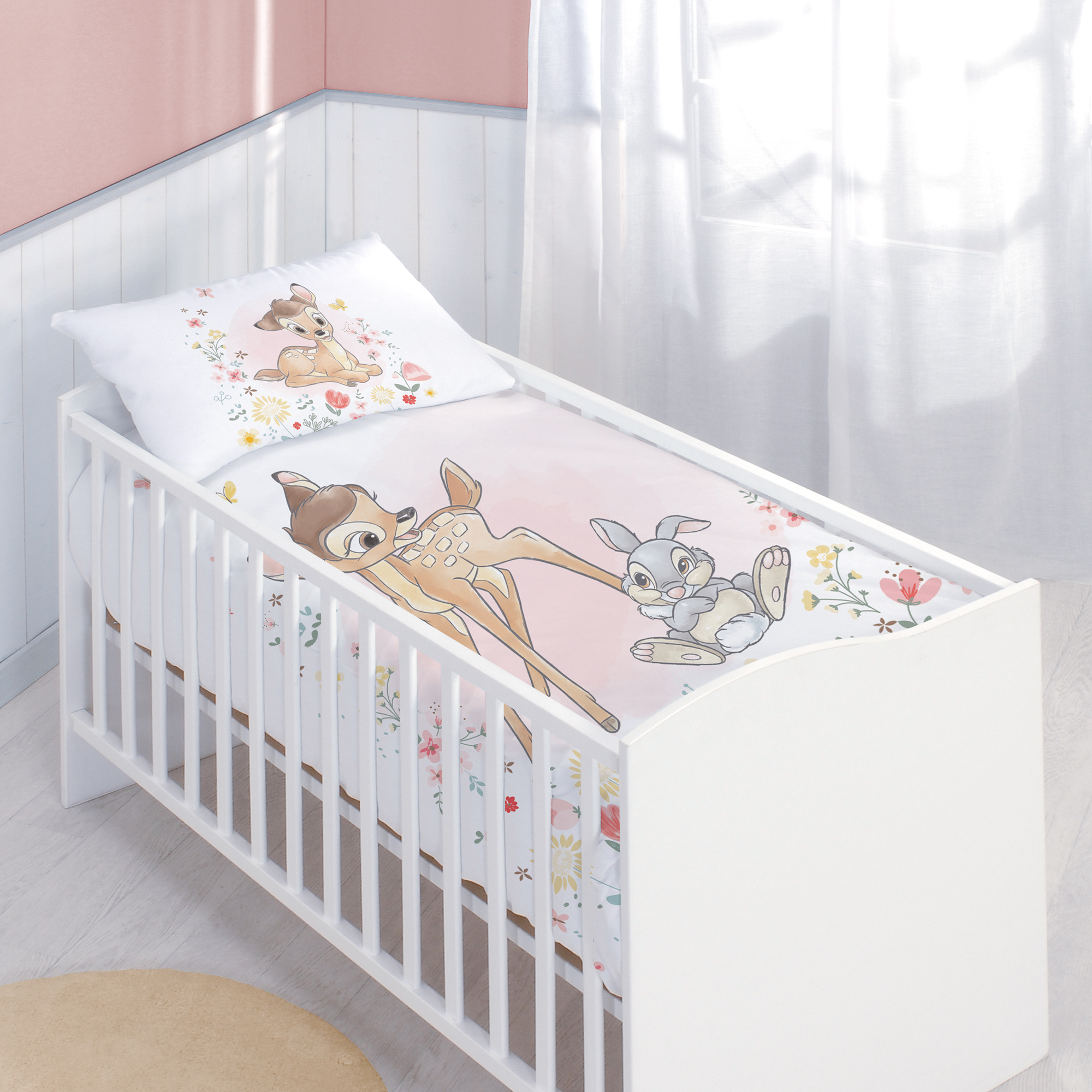 Ambiente-Aufnahme der Bambi-Babybettwäsche in einem weißen Gitterbett; der Deckenbezug zeigt Bambi und Klopfer auf einem zartrosa Hintergrund mit Blumenmustern, passend abgestimmt auf das motivgleiche Kopfkissen.