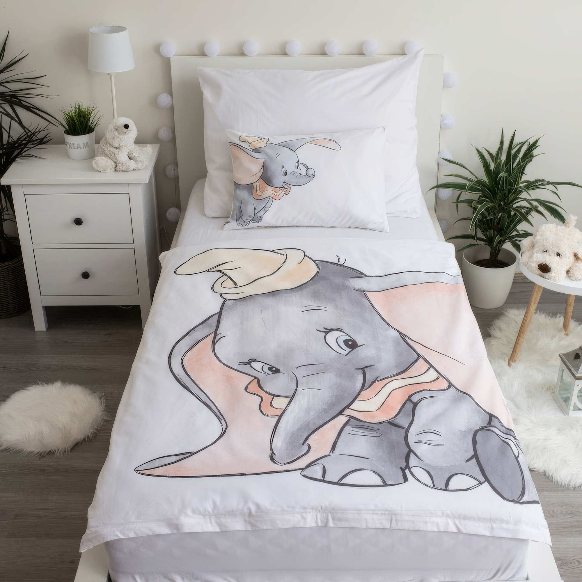 Ambiente-Aufnahme der Dumbo-Kinderbettwäsche in einem weißen Bett; der weiße Deckenbezug zeigt eine große, skizzierte Grafik des kleinen Elefanten Dumbo mit gelbem Hut, ergänzt durch ein passendes Kopfkissen.