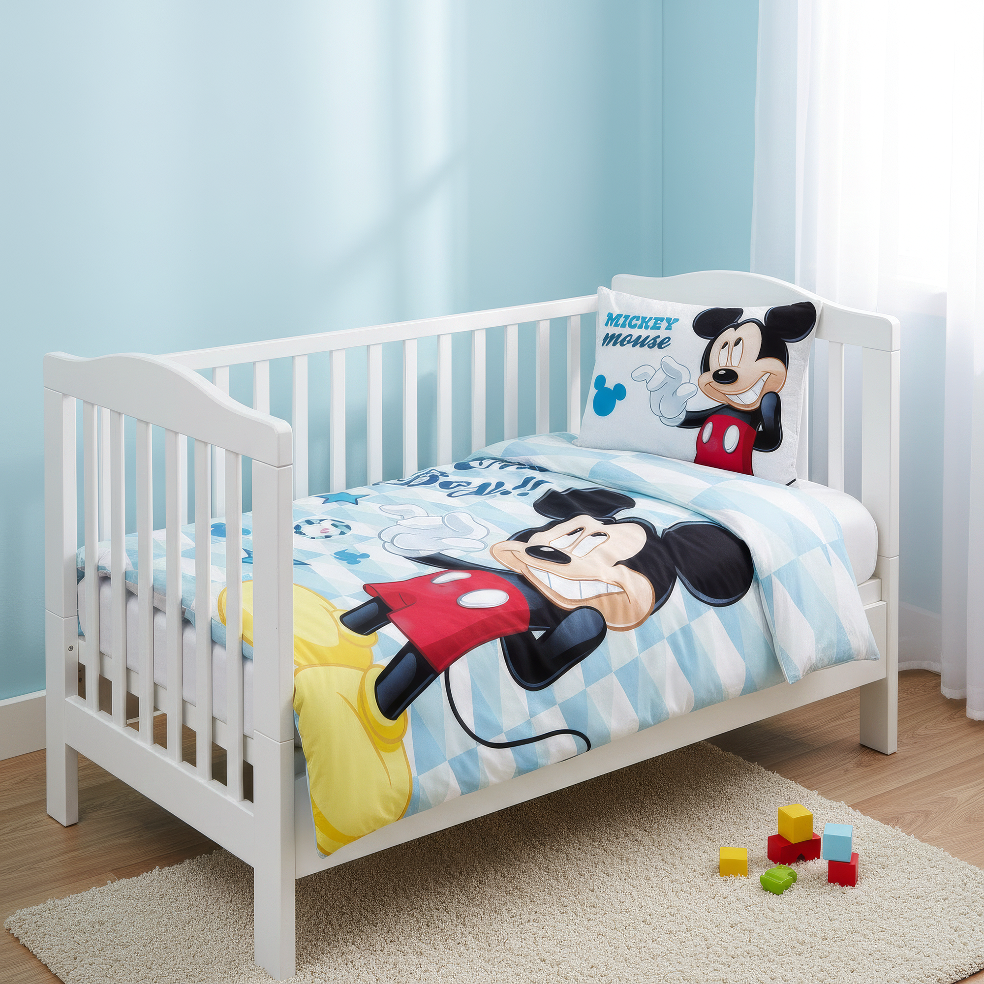 Ambiente-Aufnahme der Micky-Maus-Babybettwäsche in einem weißen Gitterbett; der hellblau karierte Deckenbezug zeigt eine große Grafik von Micky Maus, während das passende Kopfkissen ordentlich auf der Matratze liegt.
