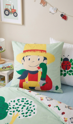 Detailaufnahme des Kopfkissens mit dem Motiv eines lächelnden Bauern im Strohhut, der eine grüne Ente im Arm hält, vor einem weiteren Kissen mit Marienkäfer-Print.
