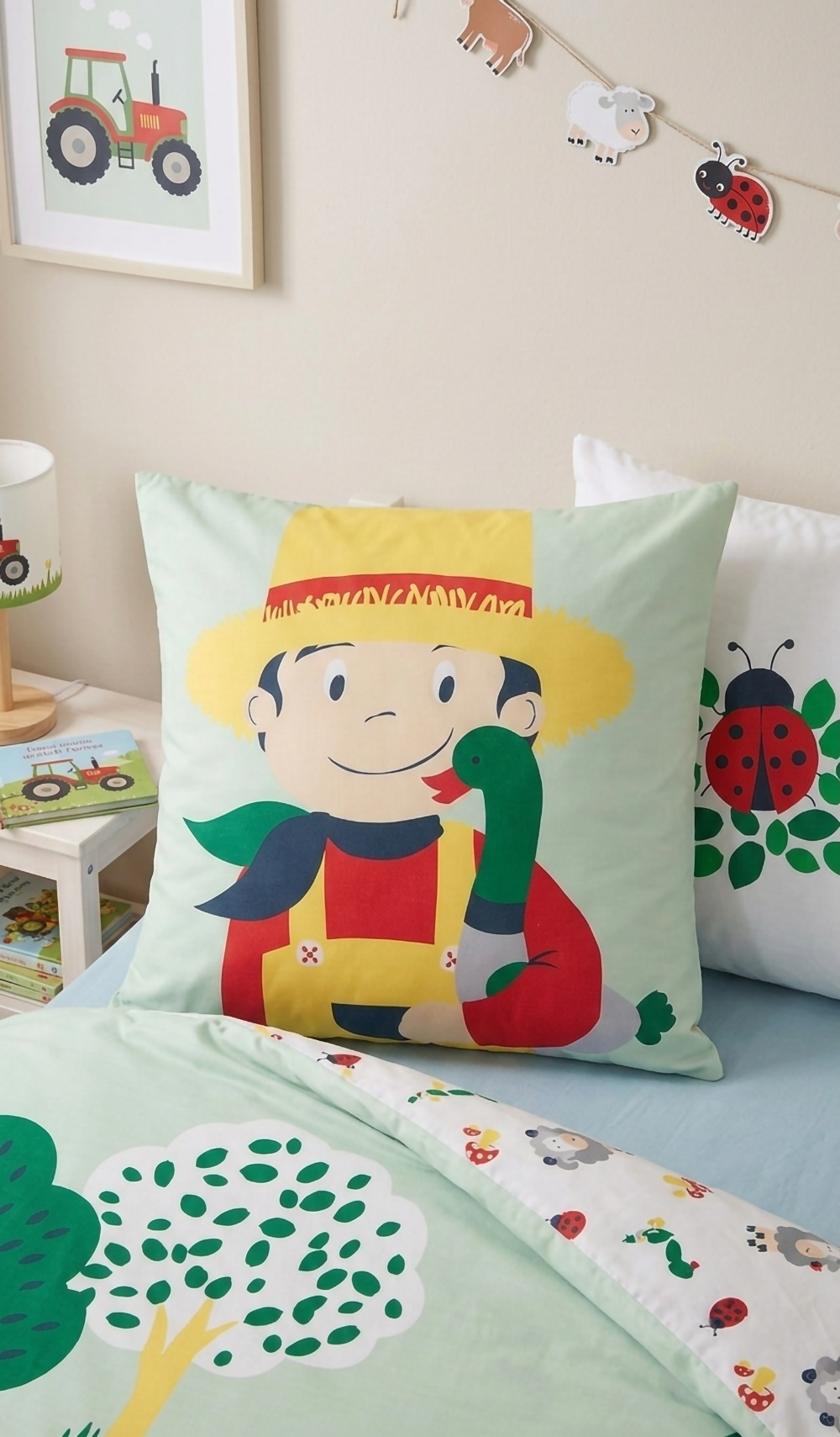 Detailaufnahme des Kopfkissens mit dem Motiv eines lächelnden Bauern im Strohhut, der eine grüne Ente im Arm hält, vor einem weiteren Kissen mit Marienkäfer-Print.