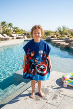 Junge trägt einen Avengers Hero Badeponcho mit Kapuze 50x115 cm, 100% Baumwolle und zeigt die Vorderseite vom Poncho. Im Hintergrund ist ein Pool.