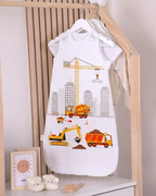 Ambiente-Aufnahme eines weißen Babyschlafsacks mit Baustellen-Motiv (Bagger, Kran), der an einer Holz-Kleiderstange hängt, dekoriert mit gestrickten Babyschuhen und einem Sonnen-Bild.