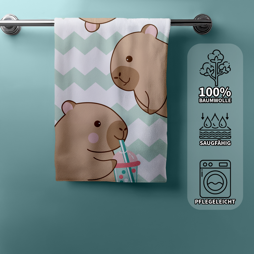 Capybara Strandtuch an einer Badetuchstange mit drei symbolischen Produkteigenschaften auf der rechten Seite: Eine Baumwollpflanze für 100% Baumwolle, Wassertropfen für saugfähig und eine Waschmaschine für pflegeleicht.