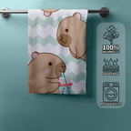 Capybara Strandtuch an einer Badetuchstange mit drei symbolischen Produkteigenschaften auf der rechten Seite: Eine Baumwollpflanze für 100% Baumwolle, Wassertropfen für saugfähig und eine Waschmaschine für pflegeleicht.