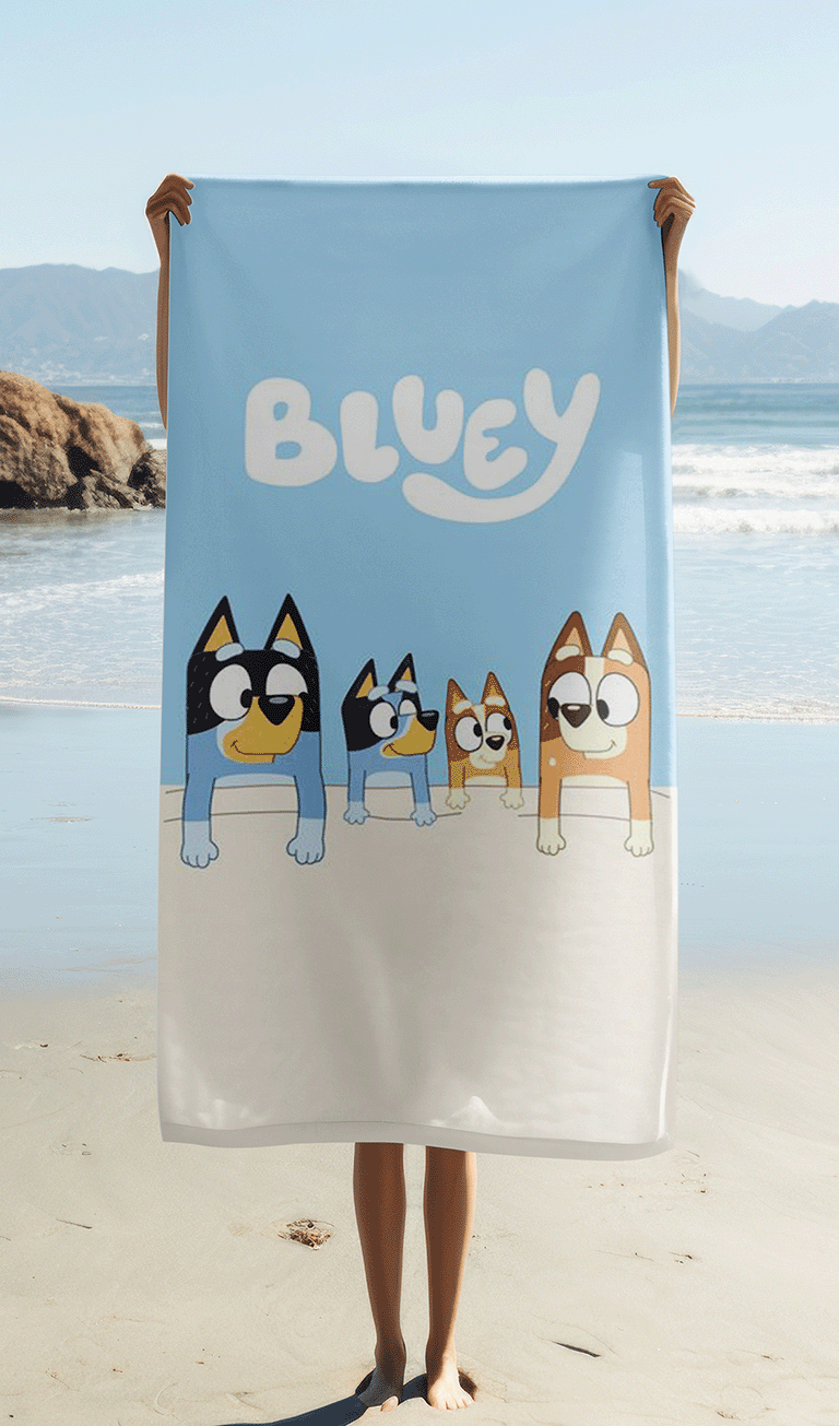 Das Bluey Strandtuch wird von einer Person am Strand hochgehalten, sodass das Motiv vollständig sichtbar ist. Im Hintergrund sind der Sandstrand, das Meer mit leichten Wellen, Felsen und eine hügelige Küstenlandschaft unter blauem Himmel zu sehen.