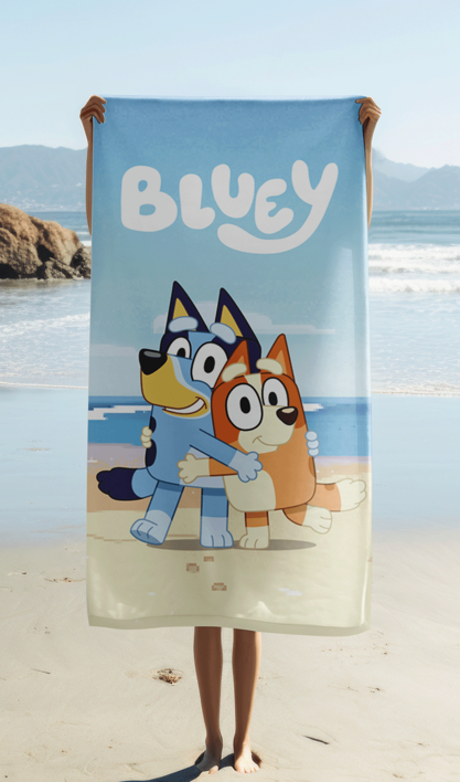Das Bluey Strandtuch wird ausgebreitet vor einer echten Meereskulisse am Sandstrand hochgehalten, sodass das Motiv mit den beiden umarmenden Charakteren komplett sichtbar ist.