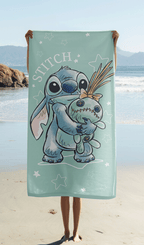 Das Stitch Strandtuch wird ausgebreitet vor einer Meereskulisse am Sandstrand hochgehalten, sodass das komplette Motiv mit Stitch und Puppe Scrump perfekt sichtbar ist.