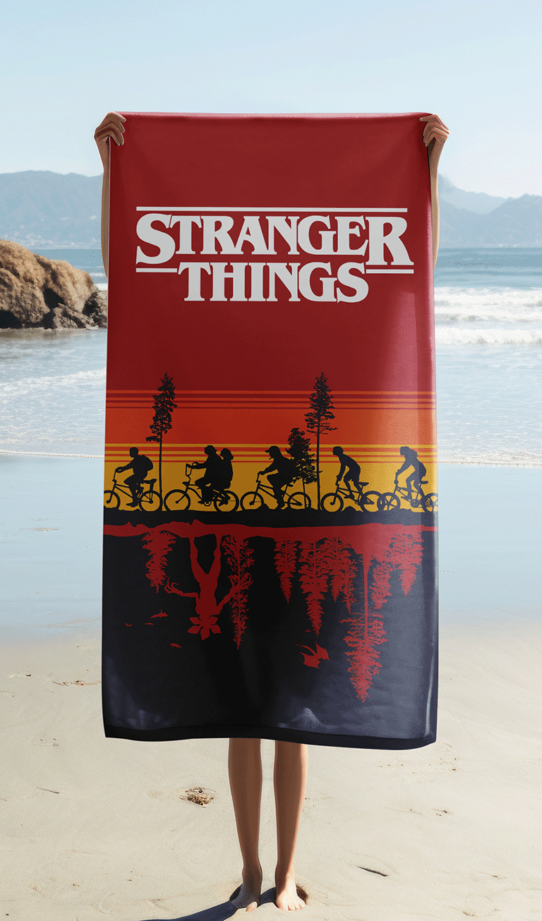 Das Stranger Things Strandtuch wird ausgebreitet vor einer Meereskulisse am Sandstrand hochgehalten, sodass das weiße Serienlogo im Zentrum komplett sichtbar ist.