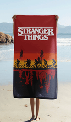 Das Stranger Things Strandtuch wird ausgebreitet vor einer Meereskulisse am Sandstrand hochgehalten, sodass das weiße Serienlogo im Zentrum komplett sichtbar ist.