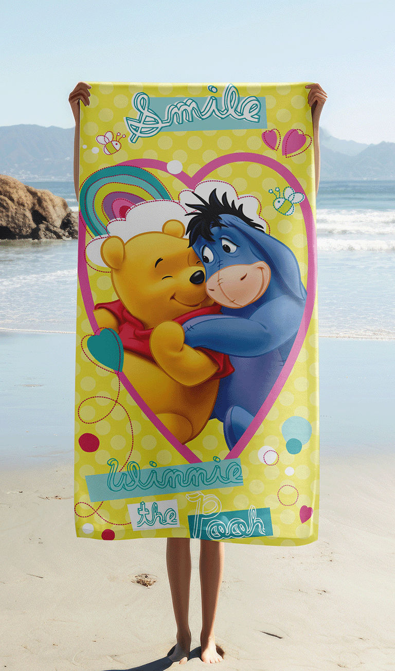 Das Winnie Puuh Strandtuch wird am Sandstrand hochgehalten, sodass das Motiv mit Puuh, I-Aah, dem rosa Herzen und dem gelben Hintergrund komplett sichtbar ist.