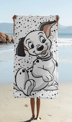Das 101 Dalmatiner Strandtuch wird am Sandstrand hochgehalten, sodass das Design mit dem Welpen Lucky auf dem gepunkteten Hintergrund komplett sichtbar ist.