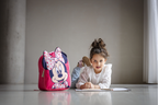 Kind liegt am Boden und zeichnet, daneben ein rosa Minnie‑Rucksack mit großem Motiv und Herzschleife.