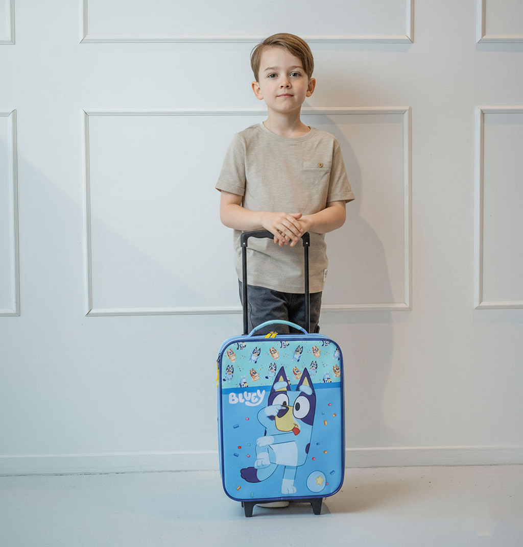 Ein kleiner Junge im beigen T-Shirt steht lächelnd hinter dem blauen Bluey Kinder-Trolleykoffer und hält den ausgezogenen Teleskopgriff.