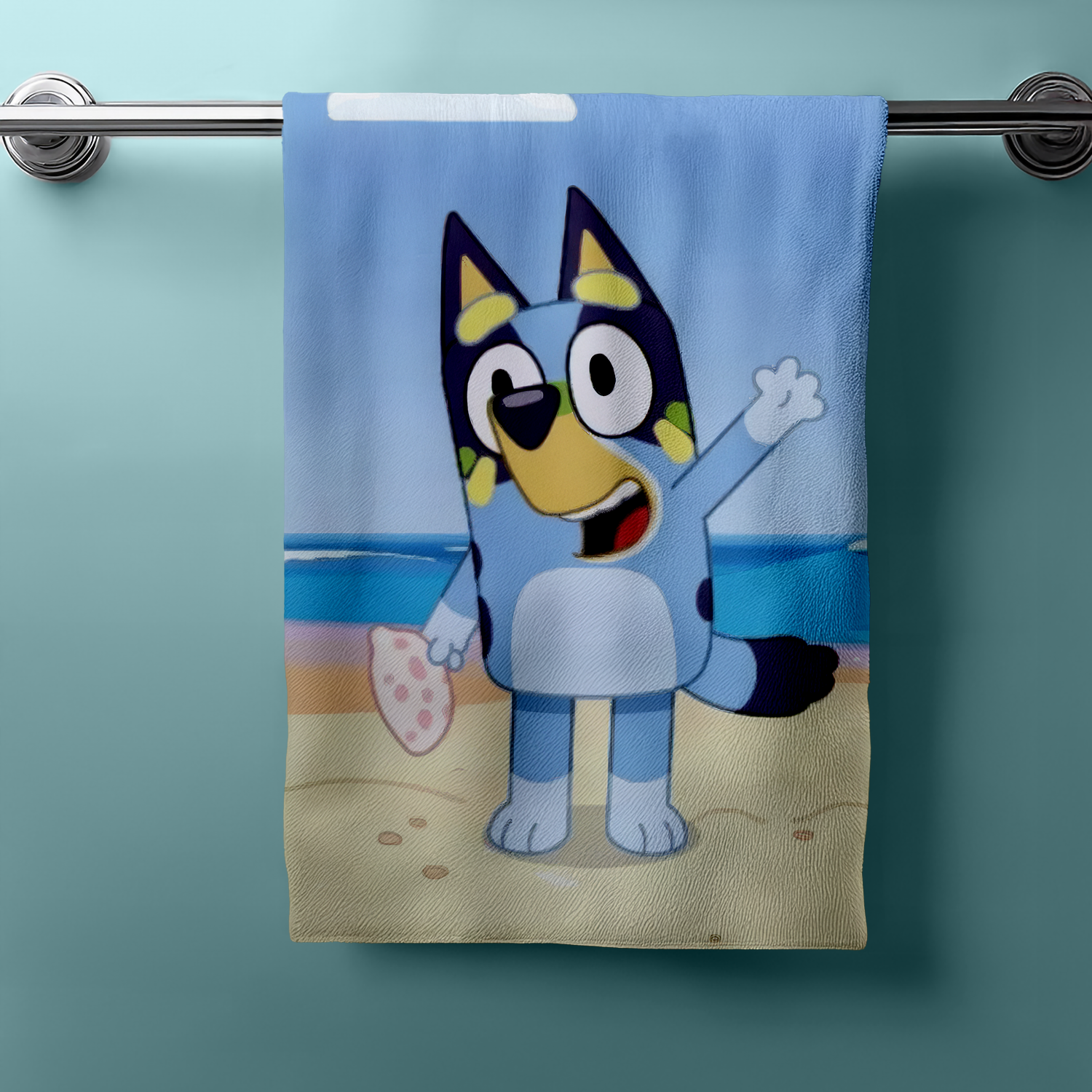 Frontansicht eines Bluey‑Handtuchs am Handtuchhalter. Bluey am Strand, winkend, mit pink gepunktetem Objekt. Heller Hintergrund, klare Farben.
