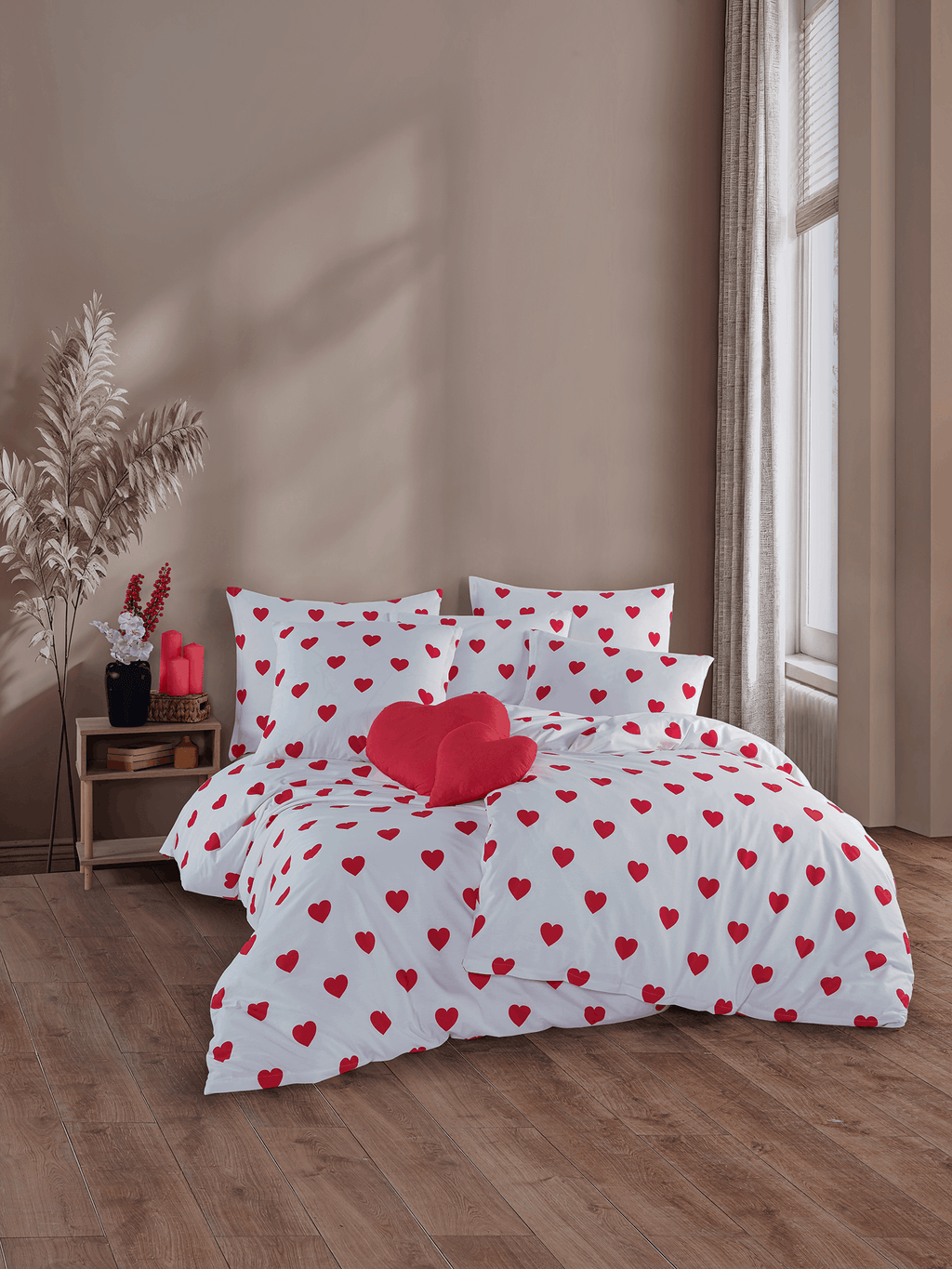Bett mit weißer Bettwäsche mit roten Herzen und rotem Herz‑Kissen. Helles Zimmer mit Holzfußboden und dekoriertem Nachttisch.