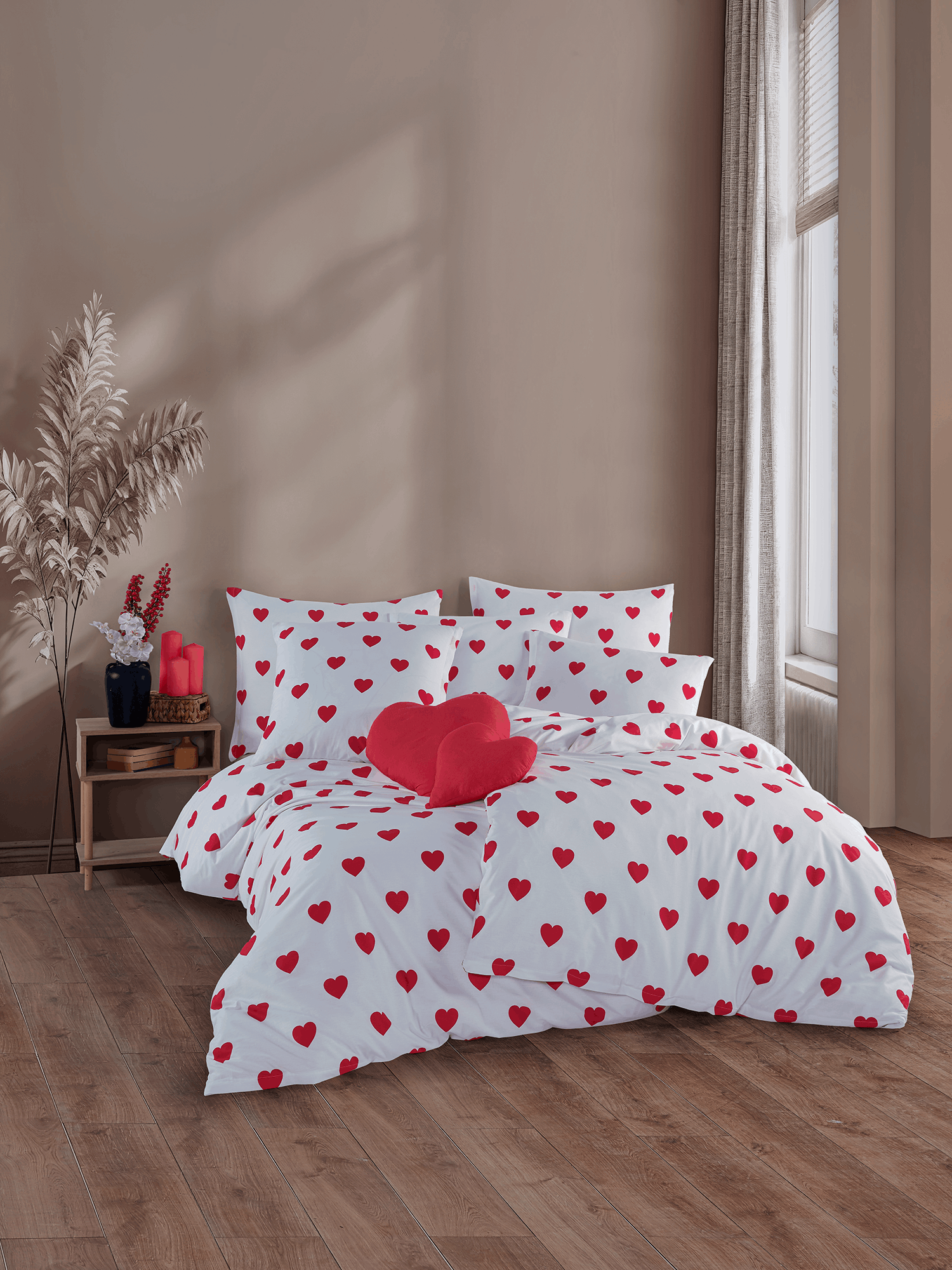Bett mit weißer Bettwäsche mit roten Herzen und rotem Herz‑Kissen. Helles Zimmer mit Holzfußboden und dekoriertem Nachttisch.
