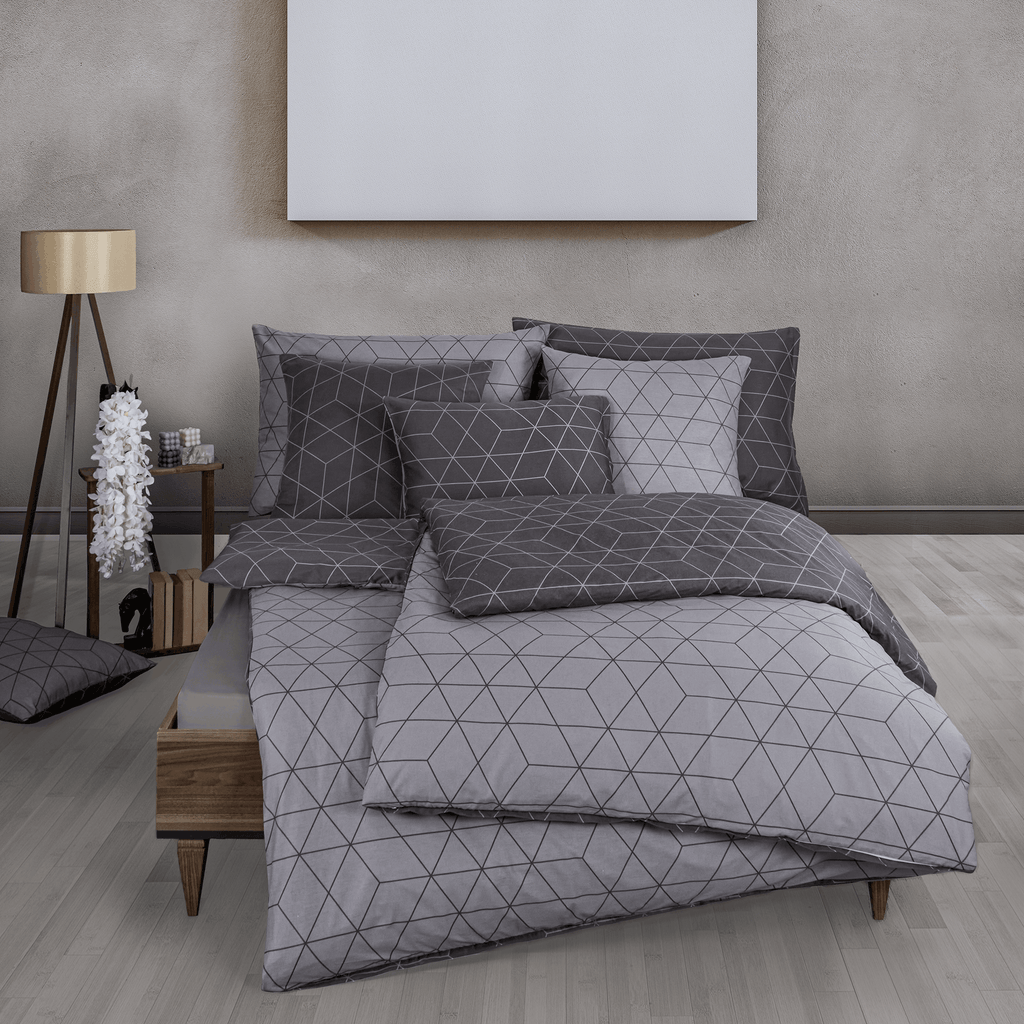 Bett mit grauer Bettwäsche im geometrischen Linienmuster. Modernes Zimmer mit hellem Holzfußboden, Nachttisch, Lampe und passendem Deko‑Kissen.