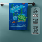 Hängendes Minecraft‑Handtuch mit Delfin, Schildkröte und Fischen. Rechts Icons: 100% Baumwolle, saugfähig, pflegeleicht.