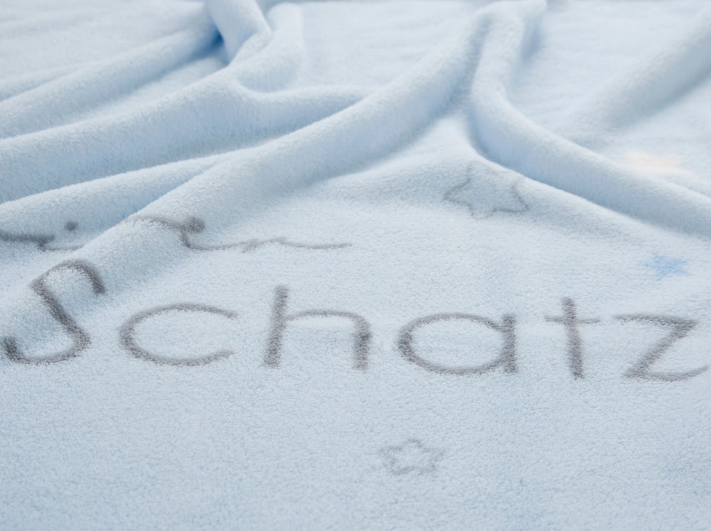Detailansicht der hellblauen Kuscheldecke mit dem Fokus auf dem grauen Schriftzug „Schatz“ und kleinen Sternen.