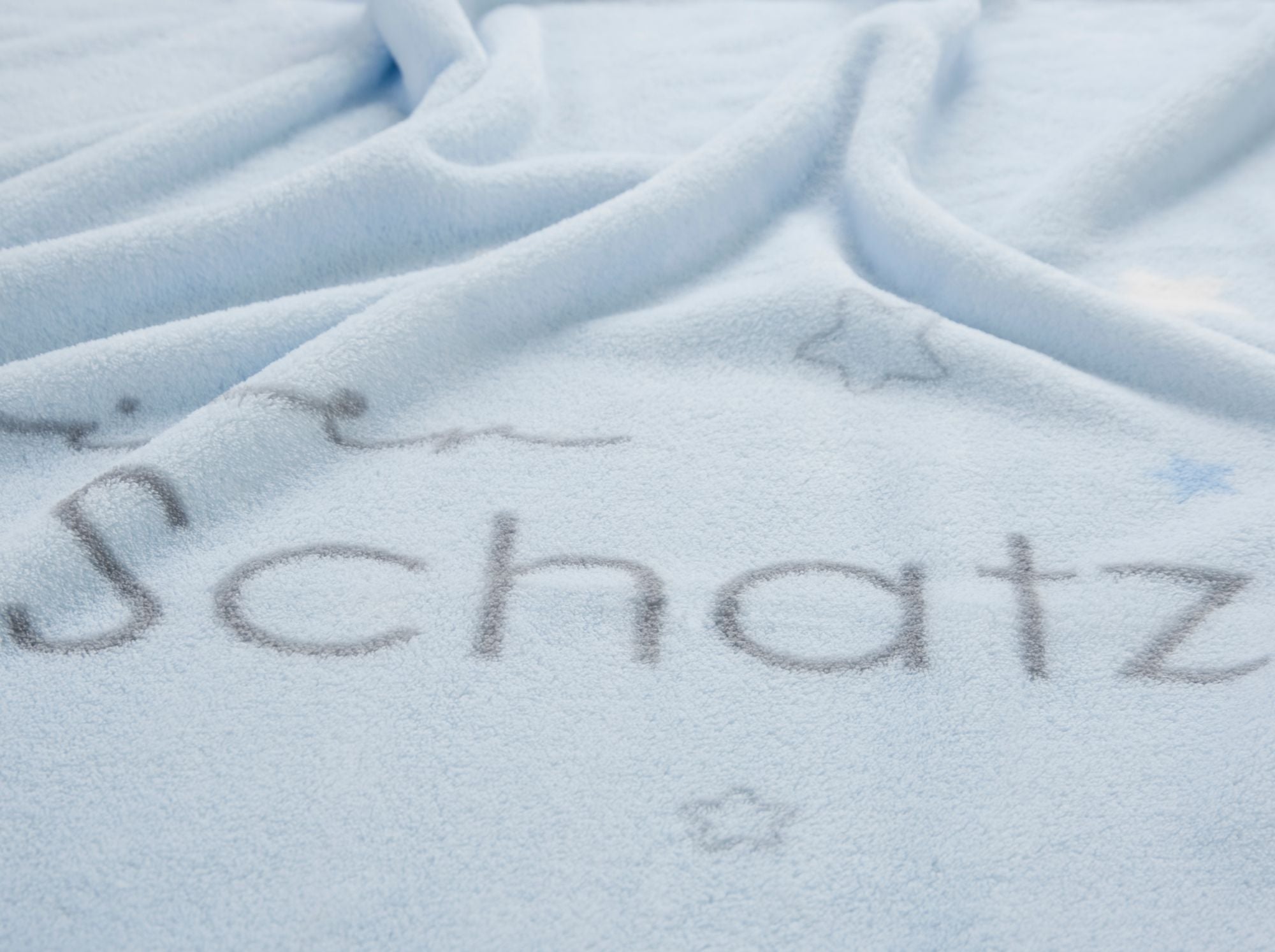 Detailansicht der hellblauen Kuscheldecke mit dem Fokus auf dem grauen Schriftzug „Schatz“ und kleinen Sternen.