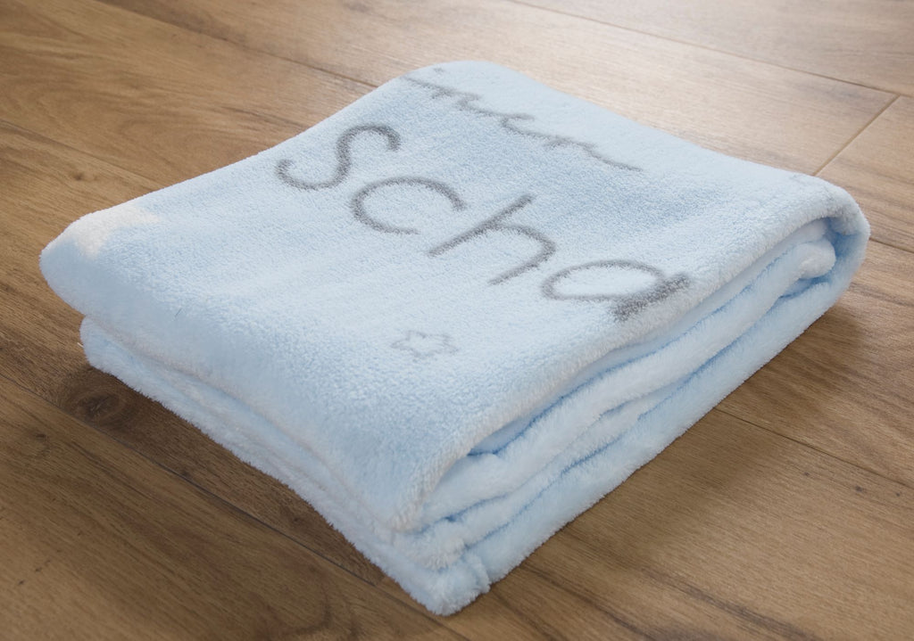 Hellblaue Babydecke zusammengelegt auf Holzboden; der graue Schriftzug „kleiner Schatz“ ist teils sichtbar.
