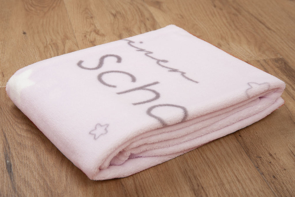 Rosa Babydecke zusammengelegt auf Holzboden; der graue Schriftzug „kleiner Schatz“ ist teils sichtbar.