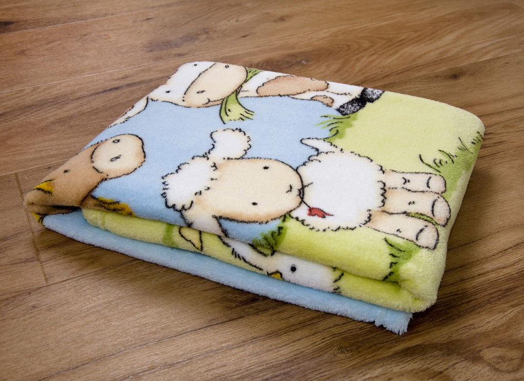 Zusammengelegte Baby-Kuscheldecke auf Holzboden; zu sehen ist ein Ausschnitt des farbenfrohen Bauernhof-Motivs mit einem Schaf, einem Pferd und einer Kuh auf einer grünen Wiese.