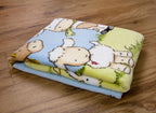Zusammengelegte Baby-Kuscheldecke auf Holzboden; zu sehen ist ein Ausschnitt des farbenfrohen Bauernhof-Motivs mit einem Schaf, einem Pferd und einer Kuh auf einer grünen Wiese.