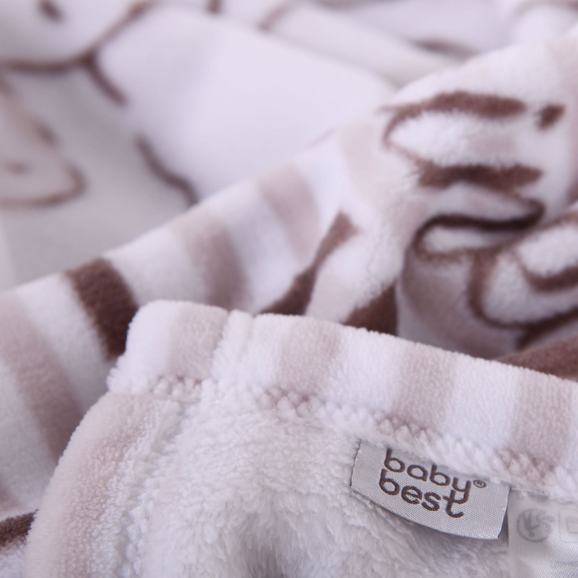 Nahaufnahme des weichen, flauschigen Materials der Kuscheldecke; im Fokus stehen der sauber verarbeitete Rand und das eingenähte „baby best“-Markenlabel vor gestreiftem Hintergrund.