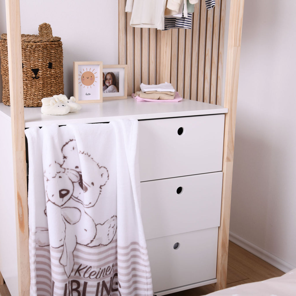 Die weiße Kuscheldecke hängt dekorativ über der Schublade einer weißen Kinderzimmerkommode; das Motiv mit Bär und Schaf sowie der Schriftzug „Kleiner Lieblingsmensch“ sind gut erkennbar.