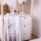Die weiße Kuscheldecke hängt dekorativ über der Schublade einer weißen Kinderzimmerkommode; das Motiv mit Bär und Schaf sowie der Schriftzug „Kleiner Lieblingsmensch“ sind gut erkennbar.