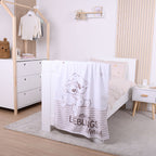 Ambiente-Aufnahme in einem hell eingerichteten Kinderzimmer; die Kuscheldecke mit dem kuschelnden Tier-Motiv hängt ausgebreitet am Fußende eines weißen Gitterbettes.
