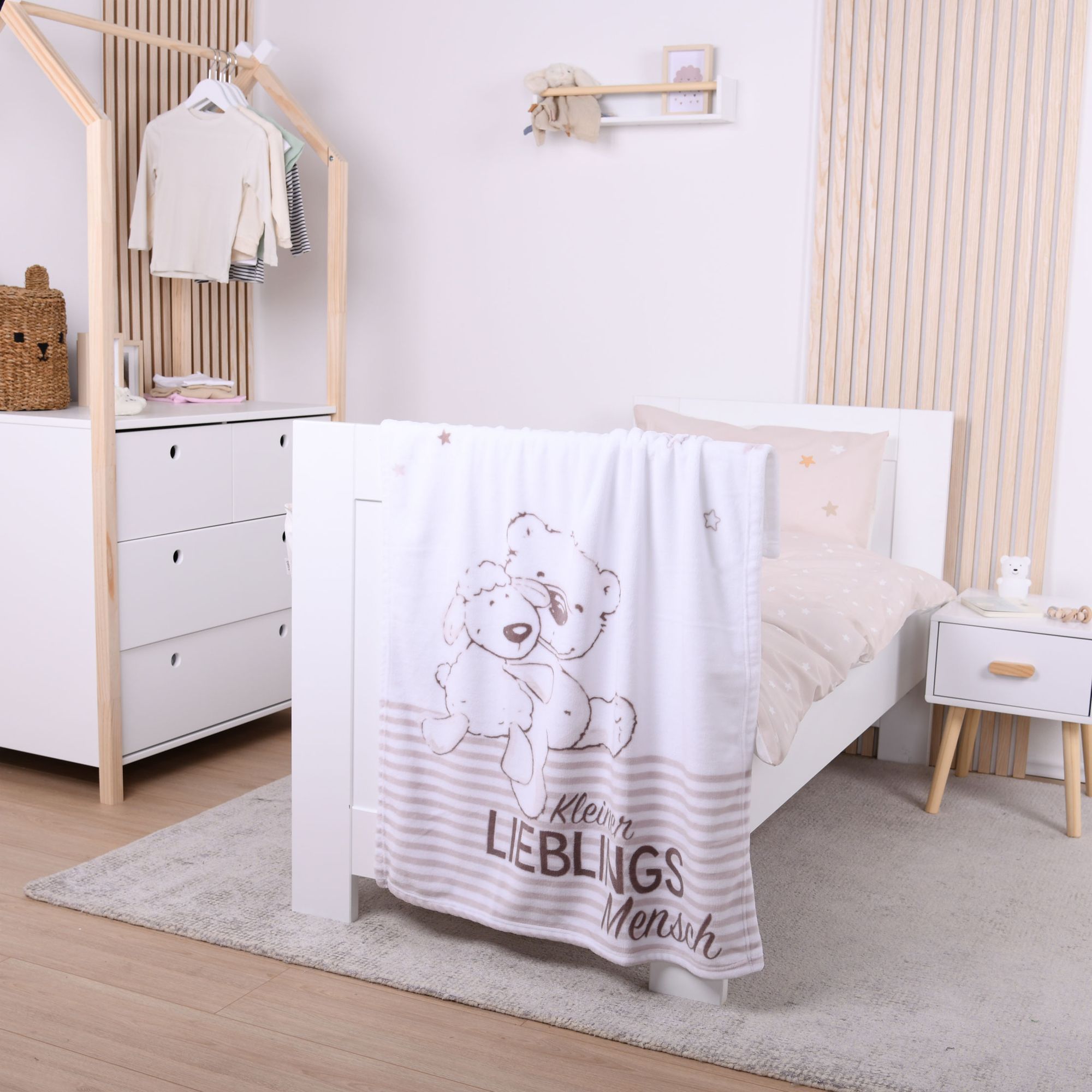 Ambiente-Aufnahme in einem hell eingerichteten Kinderzimmer; die Kuscheldecke mit dem kuschelnden Tier-Motiv hängt ausgebreitet am Fußende eines weißen Gitterbettes.