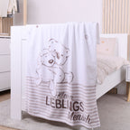 Nähere Ansicht der Kuscheldecke am weißen Kinderbett; der Fokus liegt auf dem gezeichneten Bären- und Schafsmotiv sowie dem braunen Schriftzug „Kleiner Lieblingsmensch“ über dem gestreiften Abschluss.