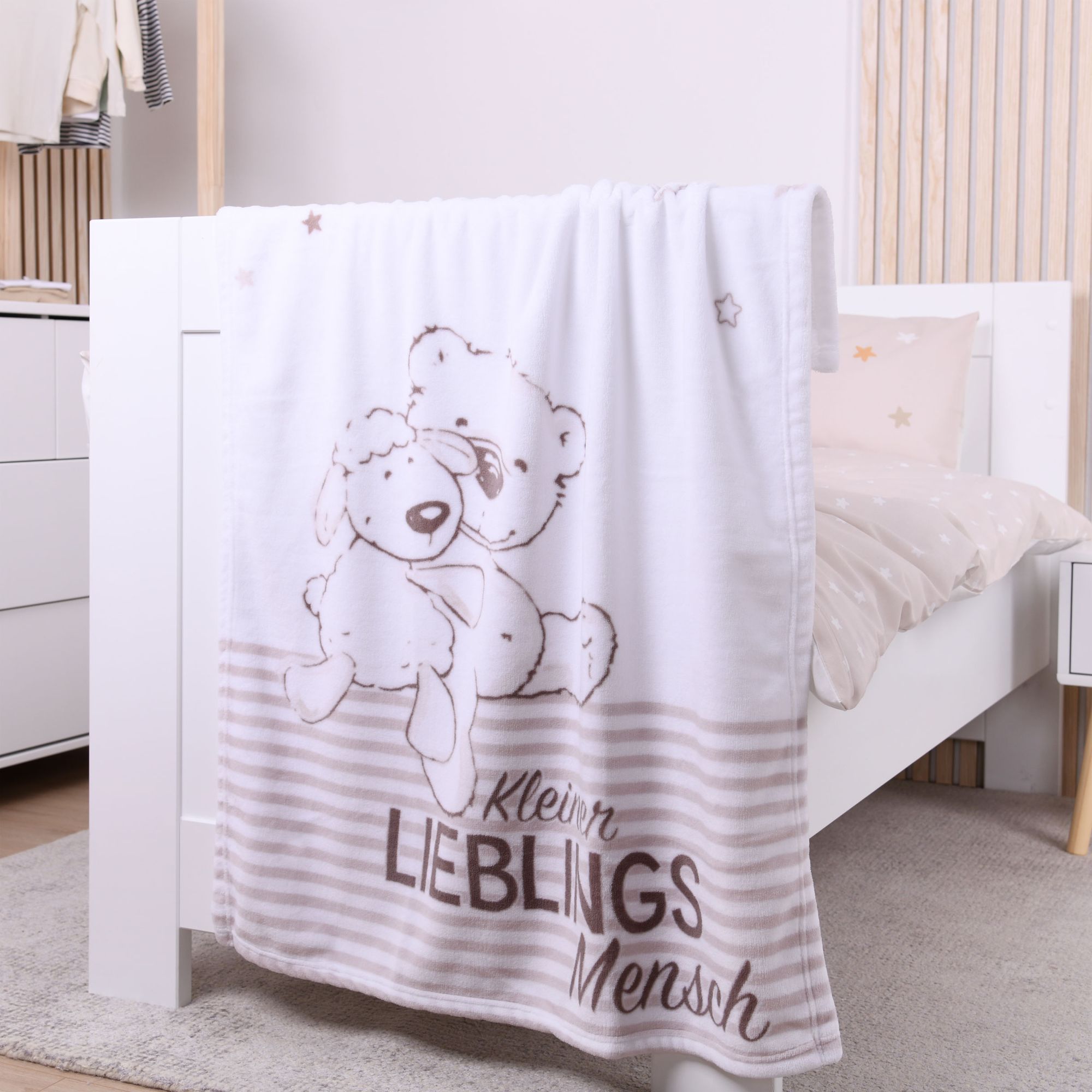 Nähere Ansicht der Kuscheldecke am weißen Kinderbett; der Fokus liegt auf dem gezeichneten Bären- und Schafsmotiv sowie dem braunen Schriftzug „Kleiner Lieblingsmensch“ über dem gestreiften Abschluss.