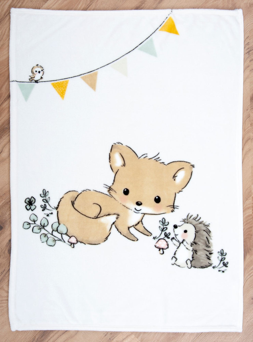 Die Baby-Kuscheldecke liegt flach ausgebreitet auf einem Holzfußboden. Das komplette Waldtiere-Motiv zeigt einen kleinen Fuchs und einen Igel auf einer Wiese mit Pilzen sowie eine farbige Wimpelkette mit einem kleinen Vogel am oberen Rand.