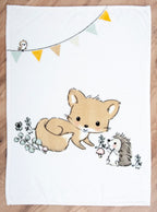 Die Baby-Kuscheldecke liegt flach ausgebreitet auf einem Holzfußboden. Das komplette Waldtiere-Motiv zeigt einen kleinen Fuchs und einen Igel auf einer Wiese mit Pilzen sowie eine farbige Wimpelkette mit einem kleinen Vogel am oberen Rand.