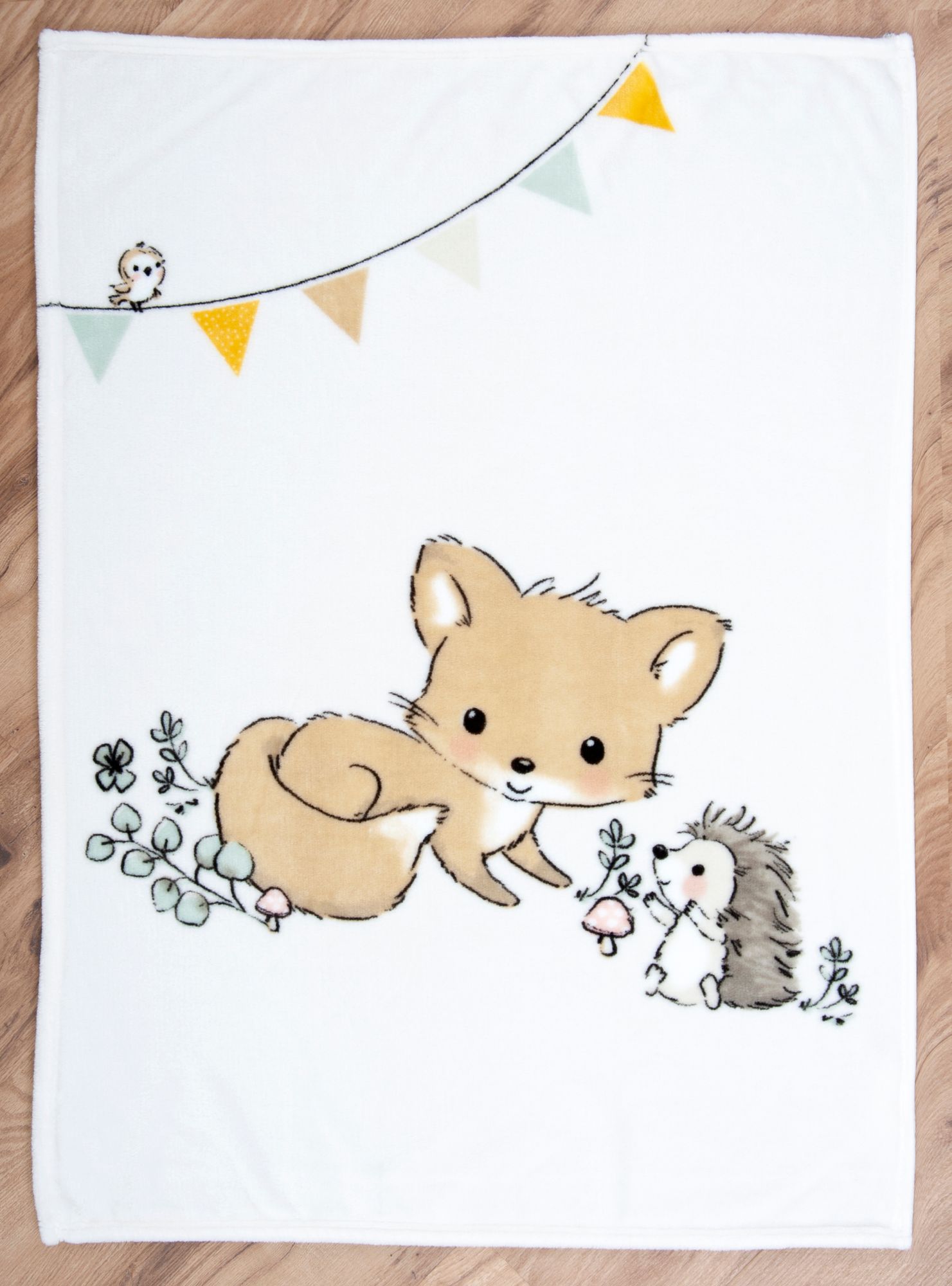 Die Baby-Kuscheldecke liegt flach ausgebreitet auf einem Holzfußboden. Das komplette Waldtiere-Motiv zeigt einen kleinen Fuchs und einen Igel auf einer Wiese mit Pilzen sowie eine farbige Wimpelkette mit einem kleinen Vogel am oberen Rand.