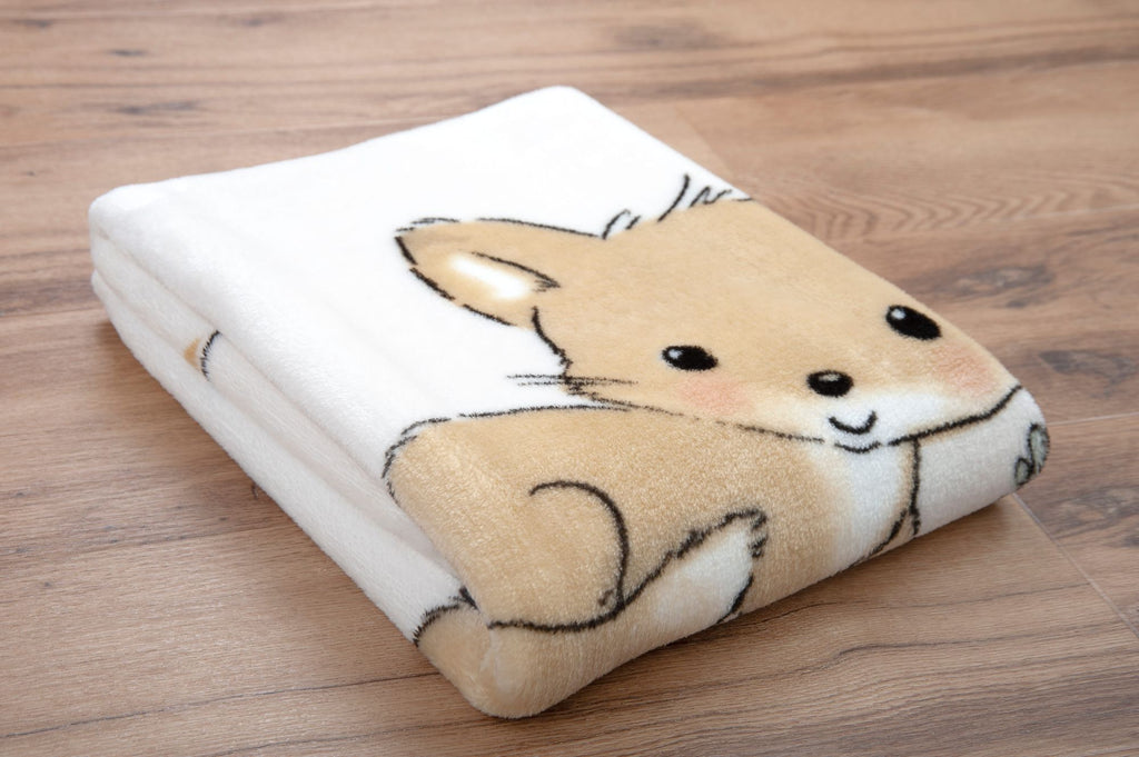 Zusammengelegte, weiße Baby-Kuscheldecke auf Holzboden; im Vordergrund ist ein Ausschnitt des niedlichen, hellbraunen Fuchs-Motivs mit großen dunklen Augen zu sehen.