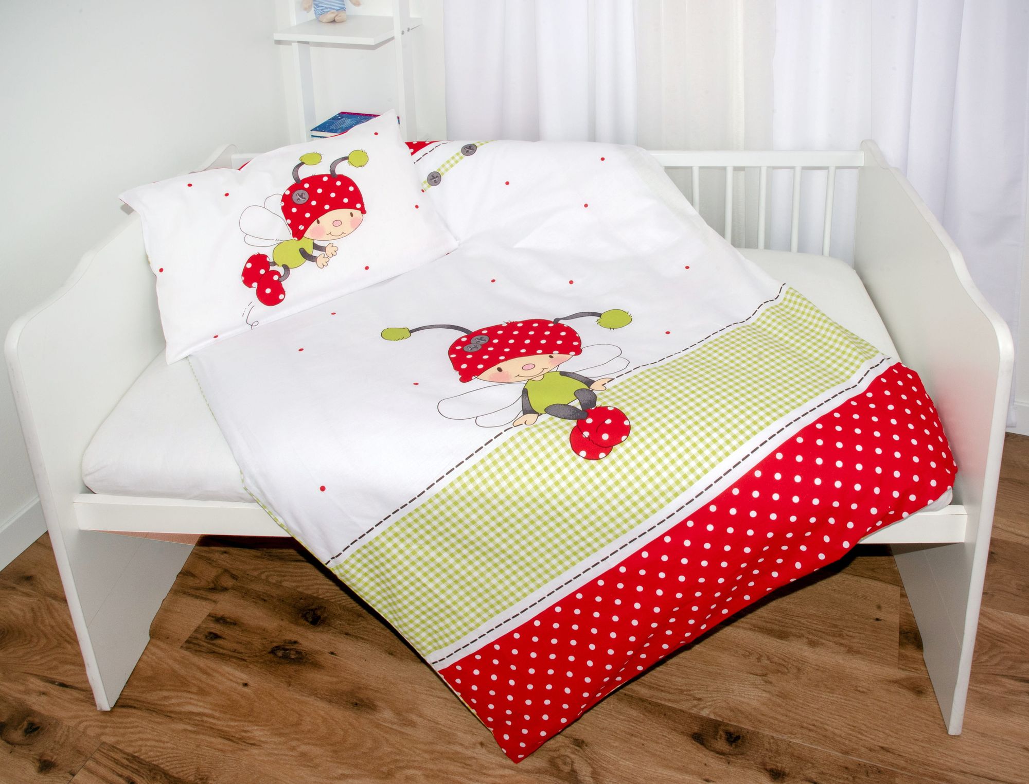 Ambiente-Aufnahme der Babybettwäsche in einem weißen Gitterbett; die Decke zeigt den sitzenden Käfer-Charakter über einem grün-weiß karierten Balken und einem rot-weiß gepunkteten Abschluss.