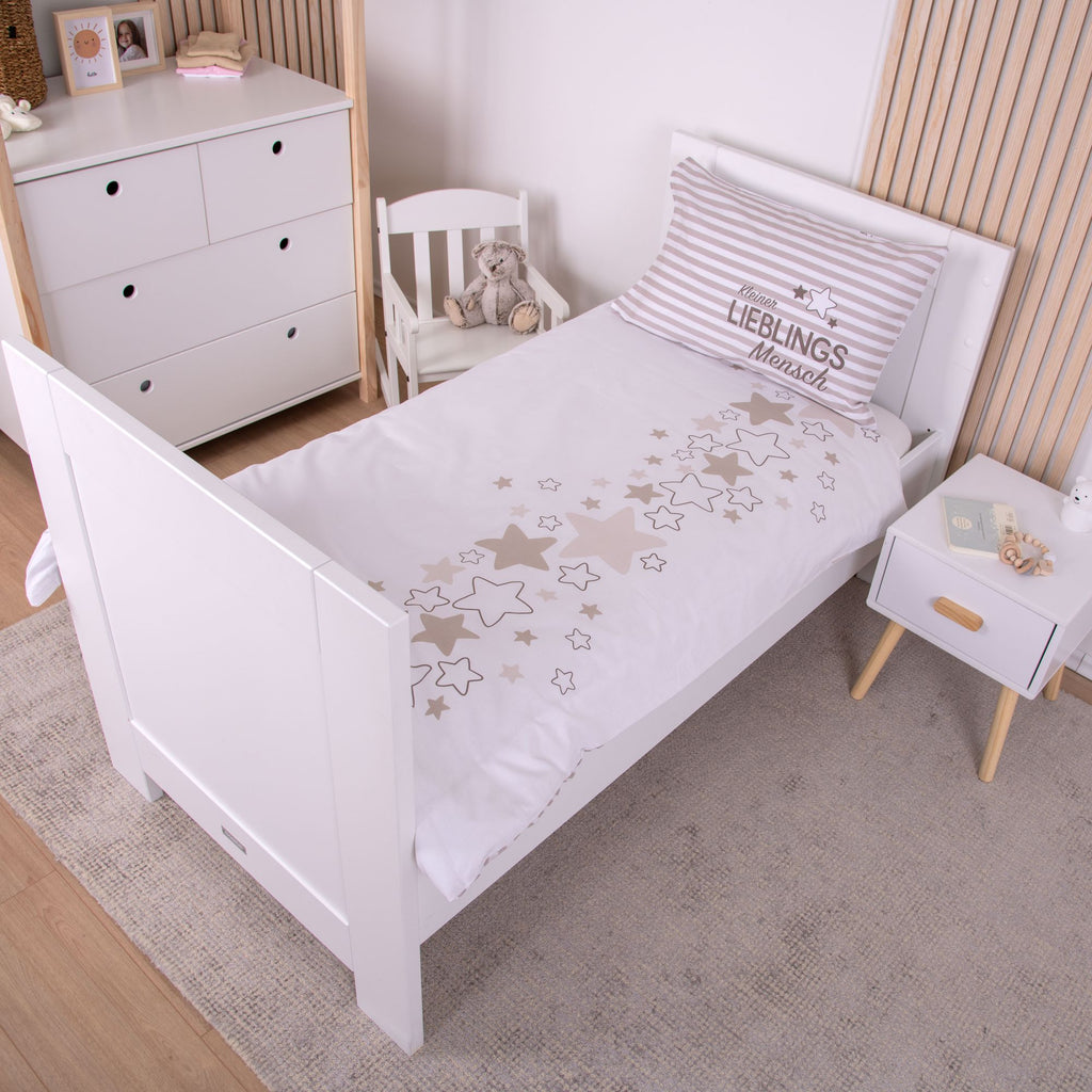 Ambiente-Aufnahme der Babybettwäsche in einem weißen Kinderbett; die Decke ist umgeschlagen und zeigt ein Design aus vielen kleinen, verstreuten Sternen, während auf dem gestreiften Kissen der Schriftzug „Kleiner LIEBLINGS Mensch“ zu sehen ist.