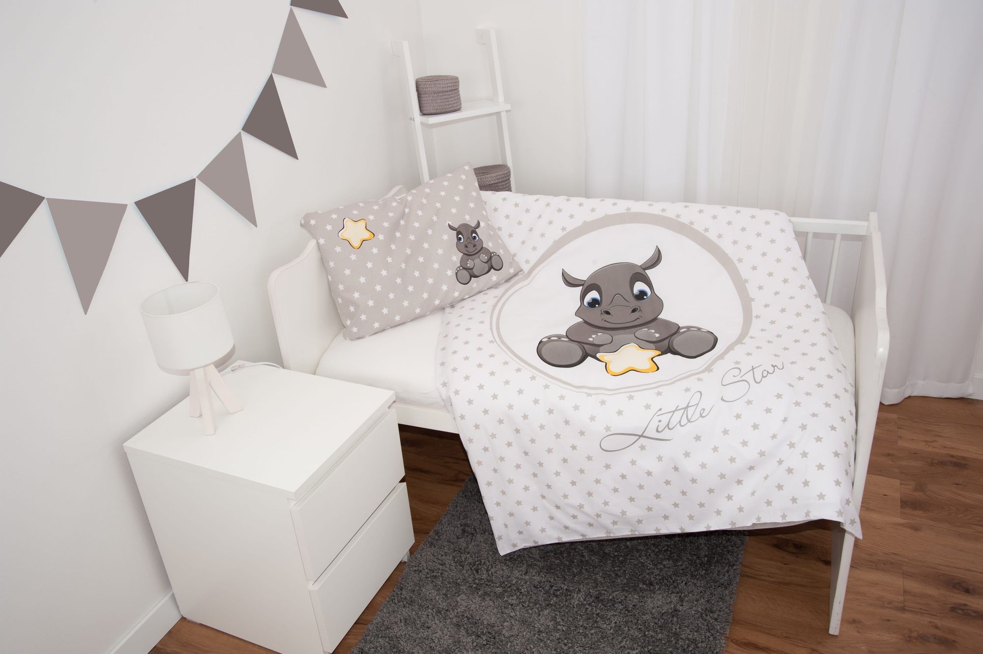 Ambiente-Aufnahme der Babybettwäsche in einem weißen Gitterbett; die Decke ist leicht ausgebreitet und zeigt ein schläfriges Nashorn in einem Kreis inmitten eines zarten Sternenmusters mit dem Schriftzug „Little Star“, während das Kissen daneben liegt.