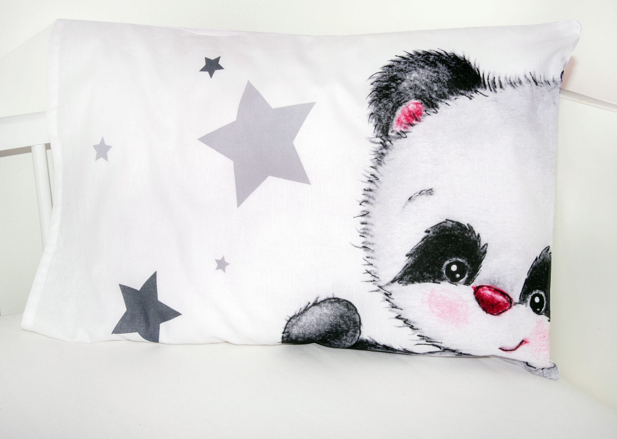 Nahaufnahme eines weißen Babykopfkissens im Gitterbett, das auf der rechten Seite das Gesicht eines süßen Pandas mit rosa Wangen und links ein Muster aus grauen Sternen zeigt.