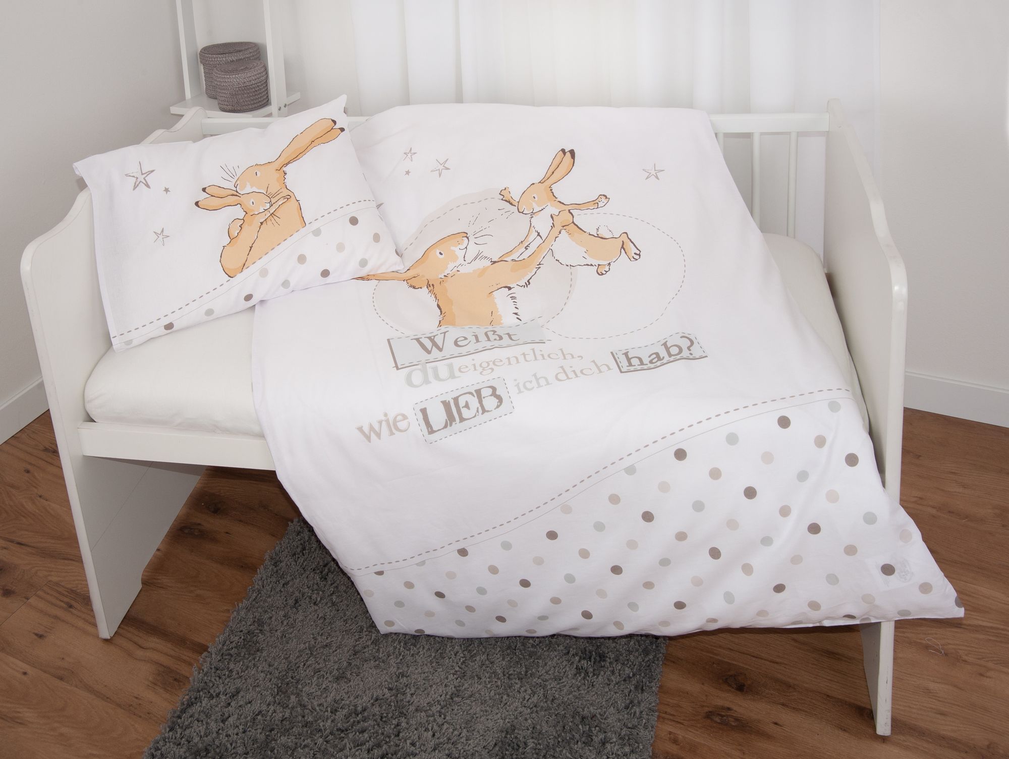 Ambiente-Aufnahme der Kinderbettwäsche in einem weißen Kinderbett; die Decke ist ausgebreitet und zeigt das Hasen-Motiv mit dem Schriftzug und einem gepunkteten unteren Rand, während das Kissen mit kuschelnden Hasen am Kopfende liegt.