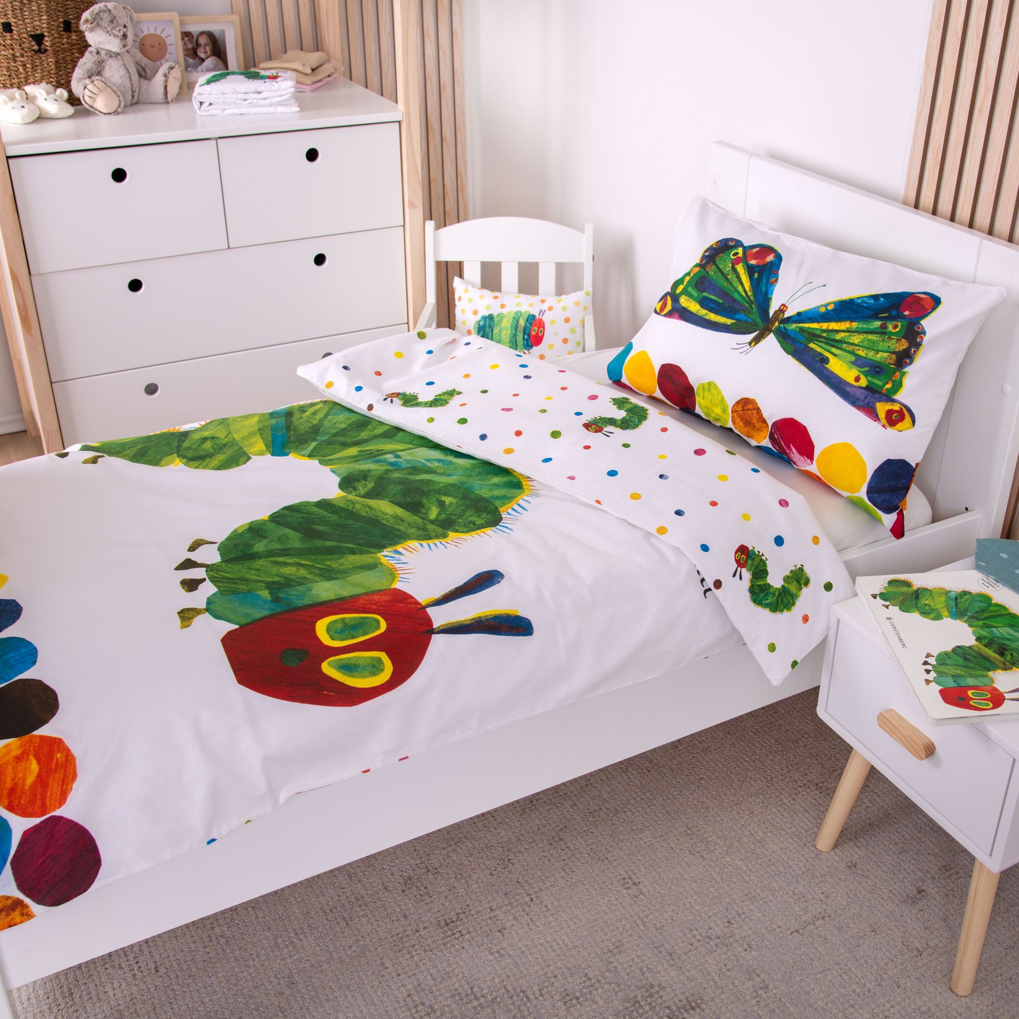 Ambiente-Aufnahme der Kinderbettwäsche „Die kleine Raupe Nimmersatt“ in einem weißen Bett; die Decke zeigt die aufgerichtete Raupe unter einer großen Sonne, während das Kissen am Kopfende mit einem bunten Schmetterling gestaltet ist.