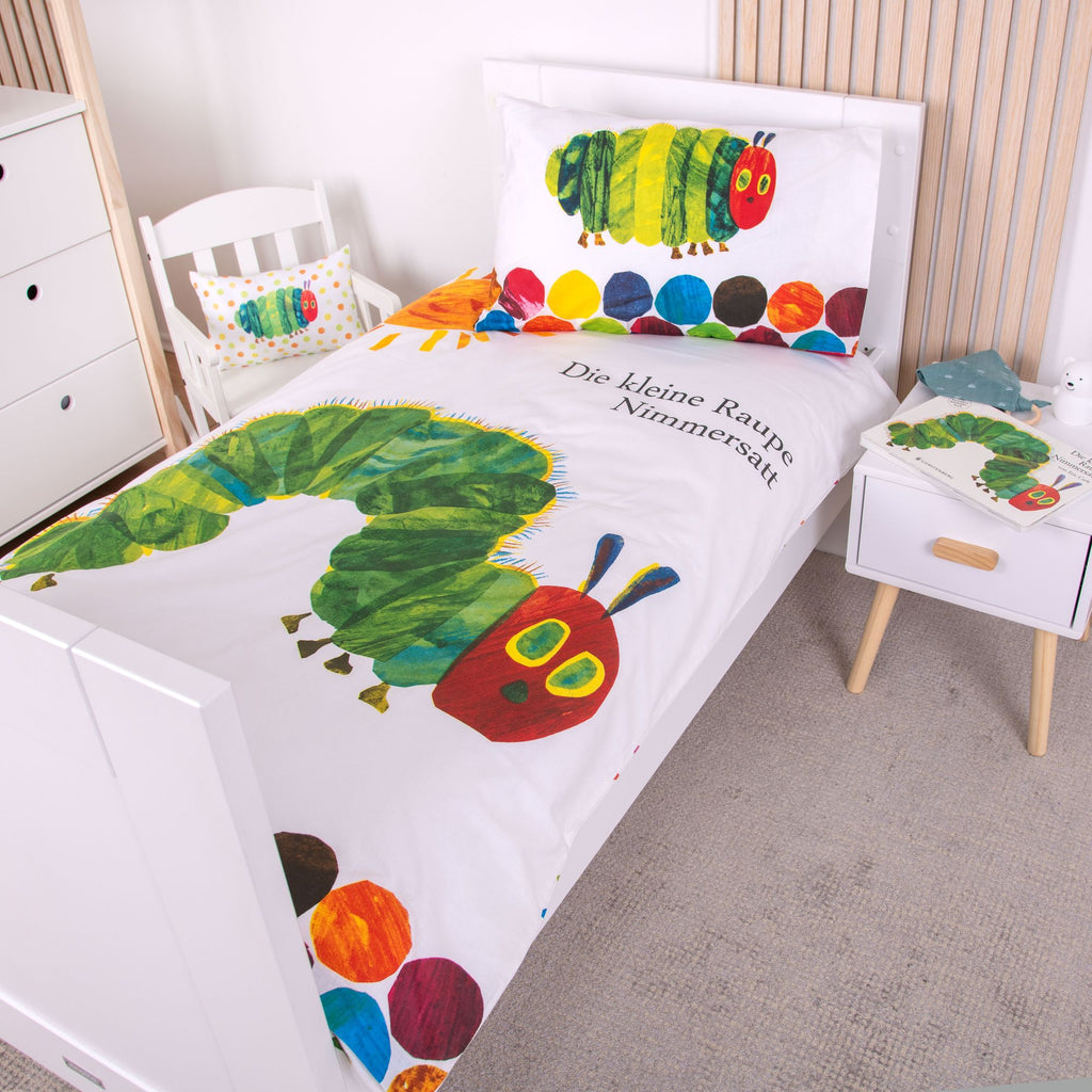 Gestreckte Ambiente-Ansicht der Bettwäsche im weißen Holzbett; zeigt den Deckenbezug mit dem Schriftzug „Die kleine Raupe Nimmersatt“ und dem großen Raupen-Motiv in voller Länge neben einem kleinen Nachttisch.