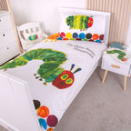 Gestreckte Ambiente-Ansicht der Bettwäsche im weißen Holzbett; zeigt den Deckenbezug mit dem Schriftzug „Die kleine Raupe Nimmersatt“ und dem großen Raupen-Motiv in voller Länge neben einem kleinen Nachttisch.