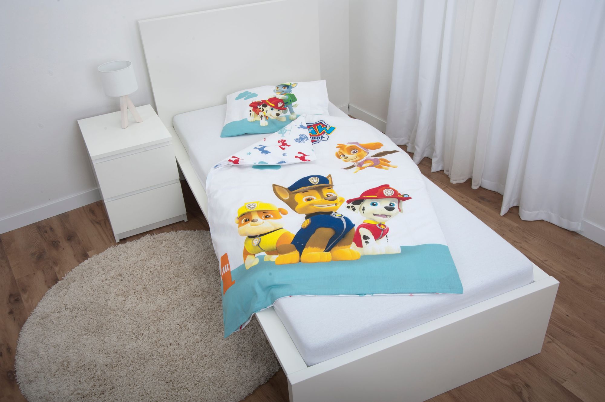 Ambiente-Aufnahme der Kinderbettwäsche in einem weißen Jugendbett; die Decke ist leicht umgeschlagen und lässt das bunte Allover-Muster der Rückseite mit kleinen Fahrzeug-Silhouetten erkennen.